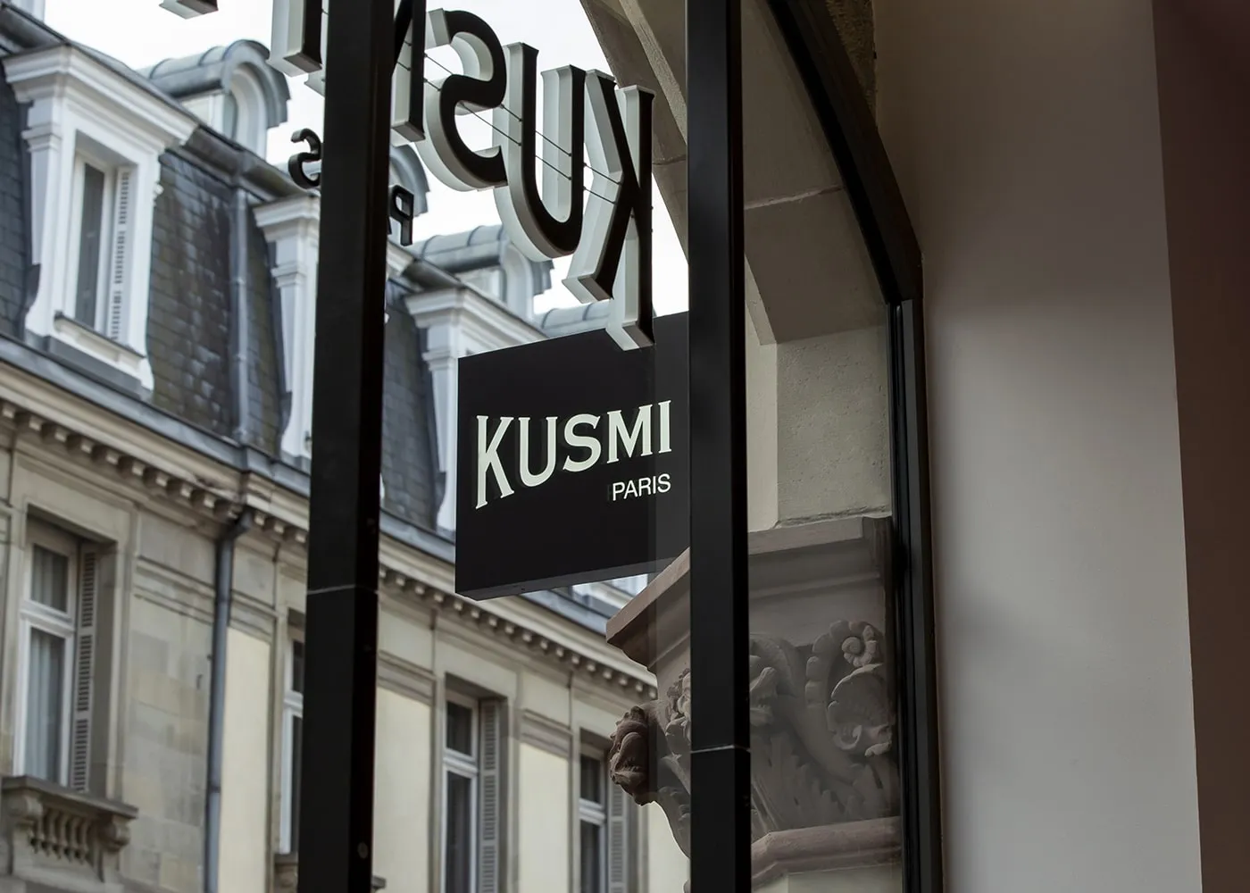 Enseigne suspendue Kusmi Paris vue depuis l’intérieur, façades haussmanniennes reflétées dans la vitrine.