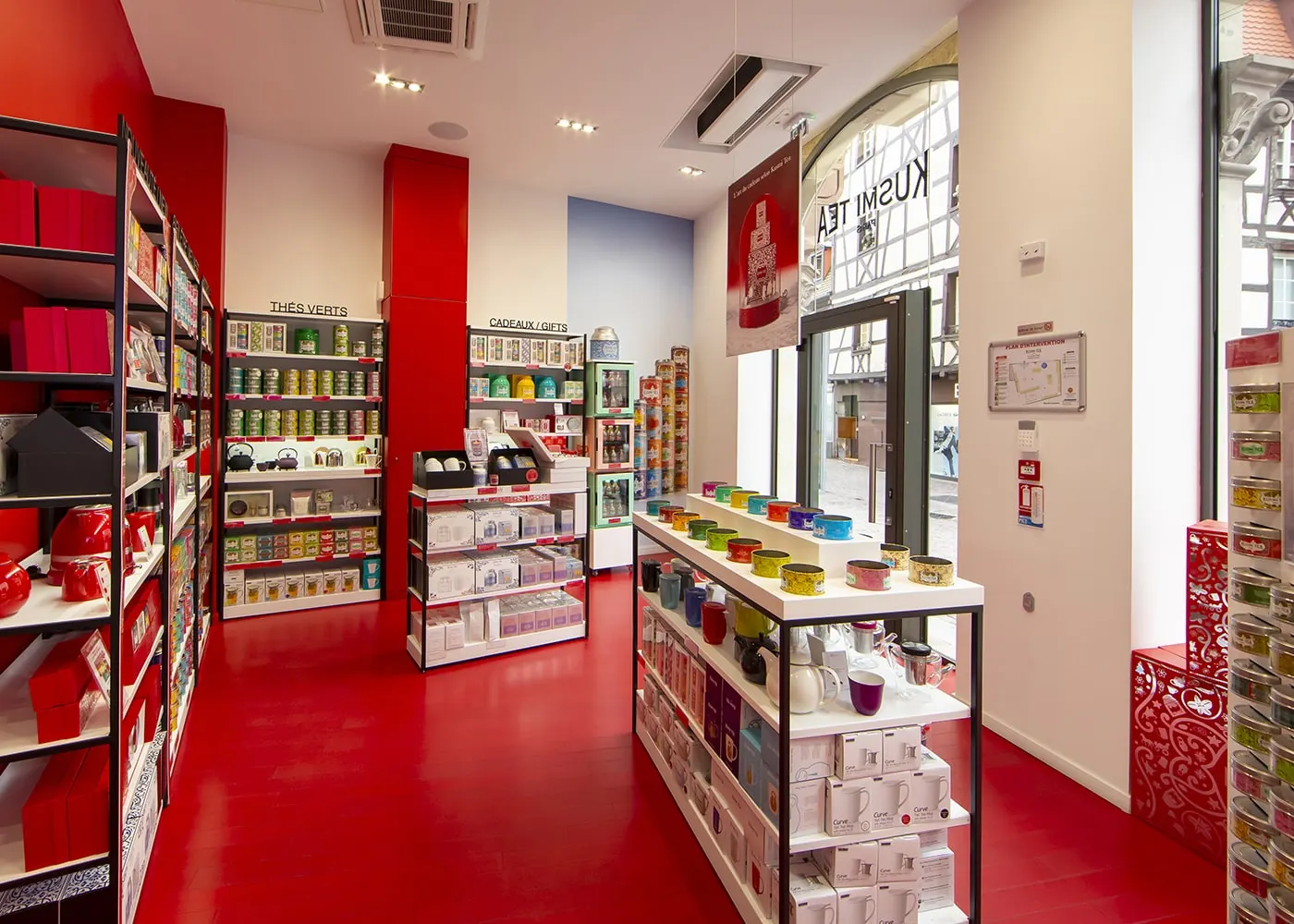 Intérieur de la boutique : sol rouge laqué, rayonnages de thés colorés et vue sur la rue par la vitrine cintrée.